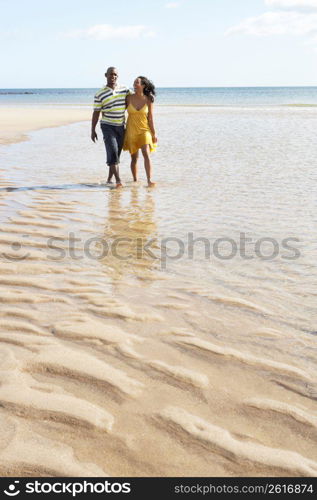 Romantic Young Couple Walking Along Shoreline Of Beach Holding Hands