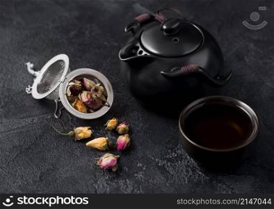 Rose buds mix tea in strainer infuser and ceramic teapot and cup on dark background.