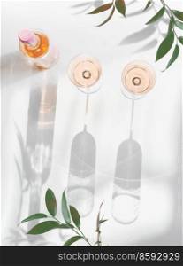 Rose wine in bottle and glasses on  white background with green branches, sunlight and shadows. Elegant modern alcohol concept. Top view