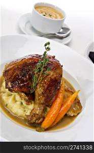 Rotisserie roasted chicken leg served over mashed potatoes with baby carrots and a gravy sauce. A fresh green sprig of rosemary tops this off. A warm bowl of soup is featured in the background.