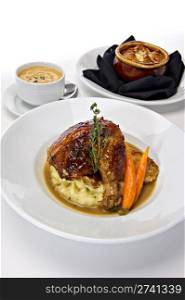Rotisserie roasted chicken leg served over mashed potatoes with baby carrots and a gravy sauce. A fresh green sprig of rosemary tops this off. Warm bowls of soup are featured in the background.