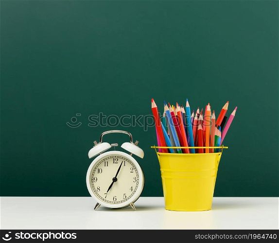 round alarm clock and multicolored pencils on the background of an empty green chalk board. Back to school