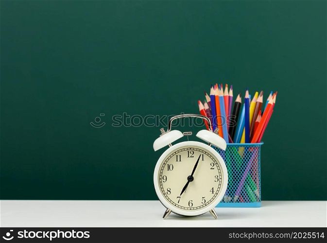 round alarm clock and multicolored pencils on the background of an empty green chalk board. Back to school