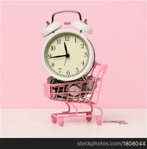 round alarm clock in a miniature shopping cart with change on a white table. Concept time is money, waste of money and poverty