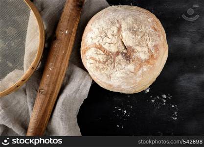 round baked bread and wooden old rolling pin on a black table, top view
