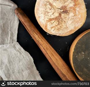 round baked bread and wooden old rolling pin on a black table, top view