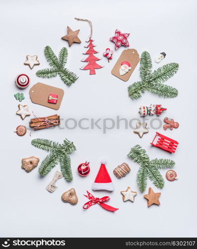 Round Christmas frame layout composition with green fir brunches, craft paper tags, holiday cookies, chocolate, Santa hat, gingerbread and red festive decoration on white background, top view.