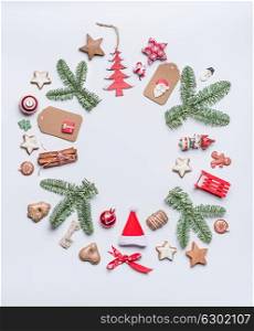 Round Christmas frame layout composition with green fir brunches, craft paper tags, holiday cookies, chocolate, Santa hat, gingerbread and red festive decoration on white background, top view.