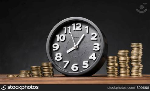 round clock between the increasing coins on wooden desk against black backdrop . Resolution and high quality beautiful photo. round clock between the increasing coins on wooden desk against black backdrop . High quality and resolution beautiful photo concept