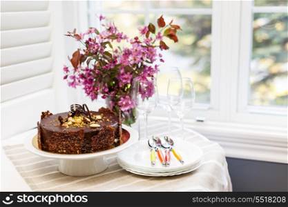 Round gourmet chocolate cake on table next to window with plates, cutlery, and pink spring bouquet