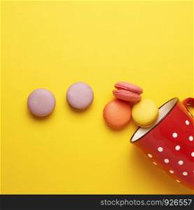 round multicolored macarons falling from a red ceramic cup with polka dots on a yellow background, top view