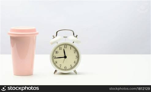 round retro alarm clock and pink ceramic cup with coffee on white table