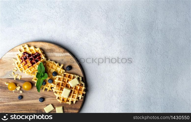 Round wooden chopping board with waffles filled with strawberry jam and blueberry with pieces of chocolate, mint and blueberry on white and grey background