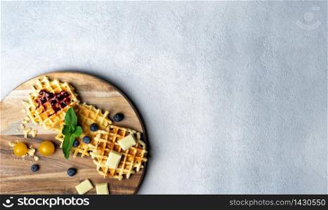 Round wooden chopping board with waffles filled with strawberry jam and blueberry with pieces of chocolate, mint and blueberry on white and grey background