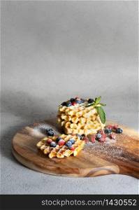Round wooden chopping board with waffles filled with strawberry jam and blueberry with mint and blueberry on white and grey background