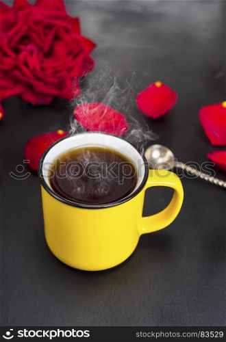 round yellow mug with hot tea on a black table, behind a red flowering rose and petals, top view