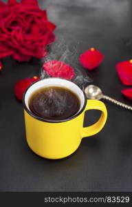 round yellow mug with hot tea on a black table, behind a red flowering rose and petals, top view