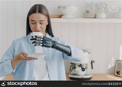 Routine of handicapped person and life quality. Pretty disabled girl is holding cup of tea with cyber hand and drinking. Concept of grasp sensors in modern robotic limb. Woman at domestic kitchen.. Routine of handicapped person. Pretty disabled girl is holding cup of tea with cyber hand.