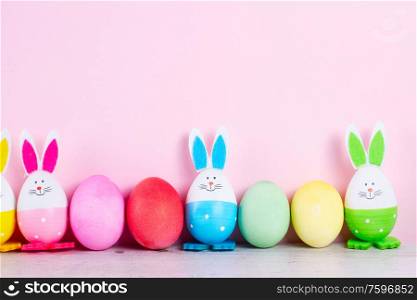 Row of funny easter bunnies and colored easter eggs on pastel pink wall background with copy space. Easter scene with colored eggs