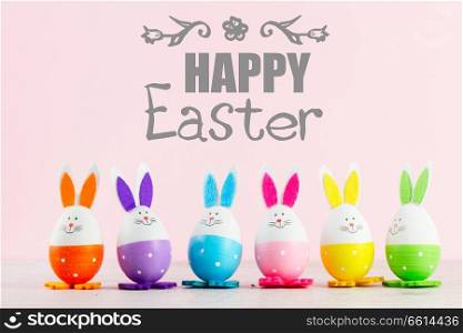 Row of funny easter bunnies of colored eggs on pink background with happy Easer greetings. Easter scene with colored eggs