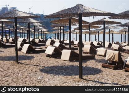 Rows of brown wooden lounge chairs and straw beach umbrellas on sea beach in the morning. Budva, Montenegro