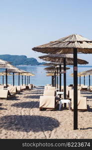 Rows of brown wooden lounge chairs and straw beach umbrellas on sea beach in the morning. Budva, Montenegro