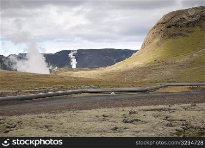 Rugged hills and mountains with steam rising, and a gas pipline along the road