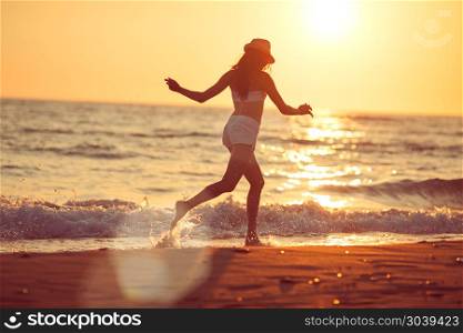 Running barefoot on the beach. Young beautiful woman running barefoot in the surf on the sand beach at sunset; lens flares