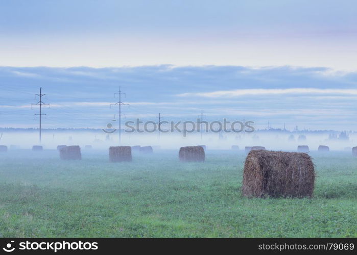 Rural landscape. Foggy field with haystacks and telegraph poles at sunset