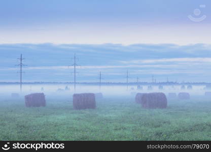 Rural landscape. Foggy field with haystacks and telegraph poles at sunset