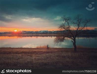 Rural landscape with sunset reflecting over the calm lake water, a bare willow tree on the dry grass meadow and a wanderer man silhouette walking a country road carrying firewood. Silent evening mood.
