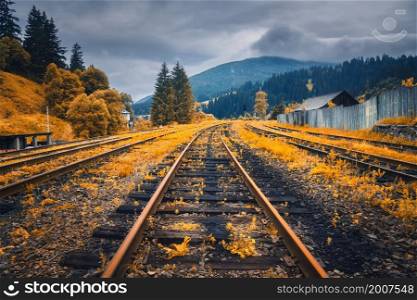 Rural railroad in mountains in overcast day in autumn. Old railway station in village at sunset. Industrial landscape with railway platform, orange trees and grass, dramatic cloudy sky, buildings
