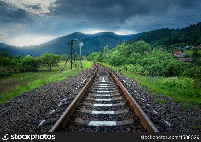 Rural railroad in mountains in overcast day in summer. Old railway station in village at sunset. Industrial landscape with railway platform, green trees and grass, dramatic cloudy sky, buildings