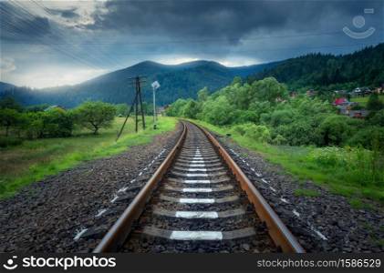 Rural railroad in mountains in overcast day in summer. Old railway station in village at sunset. Industrial landscape with railway platform, green trees and grass, dramatic cloudy sky, buildings