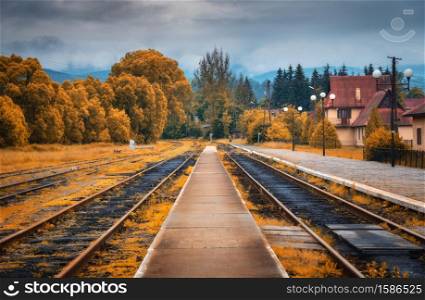 Rural railway station in autumn in cloudy day. Industrial landscape with old railway platform, orange trees, buildings, overcast sky. Railroad and beautiful forest in countryside. Railway in fall