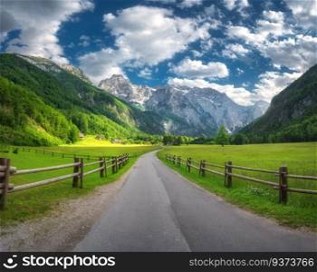 Rural road in alpine mountains, wooden fence, green meadows, trees in summer in Logar valley, Slovenia. Country road. Colorful landscape with road, rocks, field, grass, blue sky with clouds at sunset 
