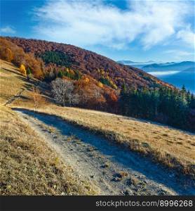Rural road in autumn mountain, colorful forest on slope and clouds between the peaks.