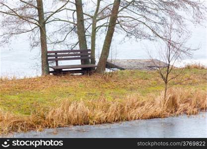 Rural scene. Single wooden bench and trees on river bank or lake shore outdoor. Autumnal tranquil landscape.