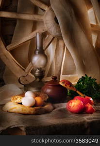 Rural still life with oil lamp, bread and vegetables, wooden wheel on background, light painting