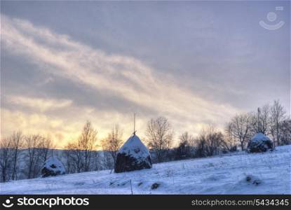 Rural winter mountain landscape with sundown and hayricks under the snow. HDR image