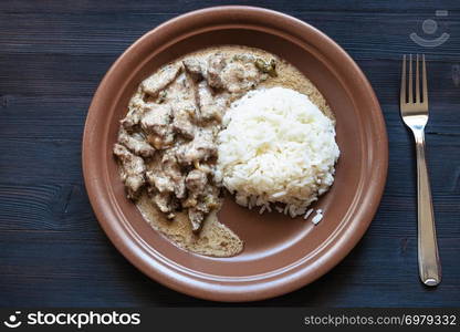 Russian cuisine dish - served portion of Beef Stroganoff (Beef Stroganov, Befstroganov) pieces of stewed meat in sour cream with boiled rice on brown plate on dark table