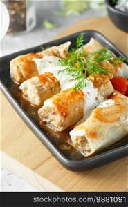 Russian stuffed cabbage rolls served with tomatoes, dill and sour cream in heat-resistant black dishes on a serving board on a gray background