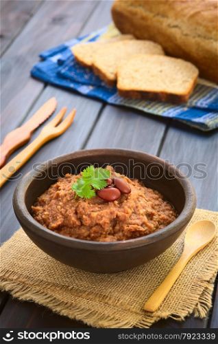 Rustic bowl of homemade red kidney bean spread garnished with kidney beans and fresh coriander leaf, wholegrain bread in the back, photographed with natural light (Selective Focus, Focus on the leaf)
