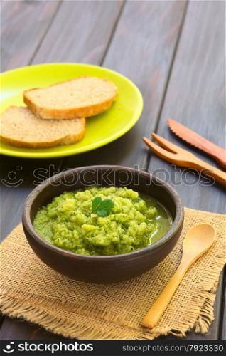 Rustic bowl of homemade zucchini and parsley spread garnished with fresh parsley leaf, slices of wholegrain bread in the back, photographed with natural light (Selective Focus, Focus on the leaf)