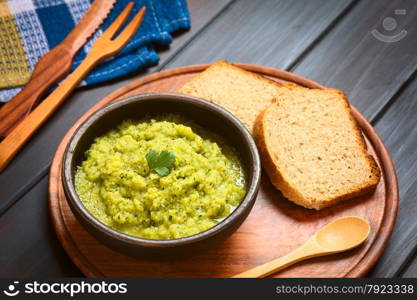 Rustic bowl of homemade zucchini and parsley spread garnished with fresh parsley leaf, slices of wholegrain bread on the side, photographed on dark wood with natural light (Selective Focus, Focus on the leaf)