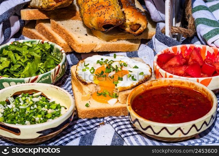 Rustic composition with sausages rolls, fried egg on toast bread, different bowls with sauce and chopped vegetables.
