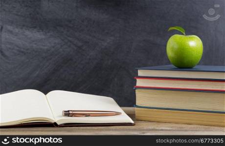 Rustic desk of student, close up view, with books, paper and pen along with a green apple in front of chalkboard. Layout in horizontal format with copy space.