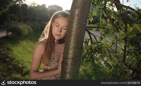 Sad attractive woman standing with head glued to tree in the park. Depressed girl with her eyes closed standing near tree outdoors. Young female suffering broken heart ache. Slow motion. Steadicam stabilized shot.
