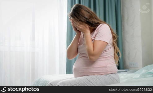 Sad crying pregnant woman suffering from depression sitting on bed and holding her head. Concept of maternal and pregnancy depression. Sad crying pregnant woman suffering from depression sitting on bed and holding her head. Concept of maternal and pregnancy depression.
