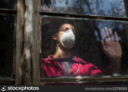 Sad depressed anxious young man wearing N95 protective medical mask looking through window,Coronavirus COVID-19 pandemic,self isolation quarantine,social physical distance,stay at home safe,hope faith